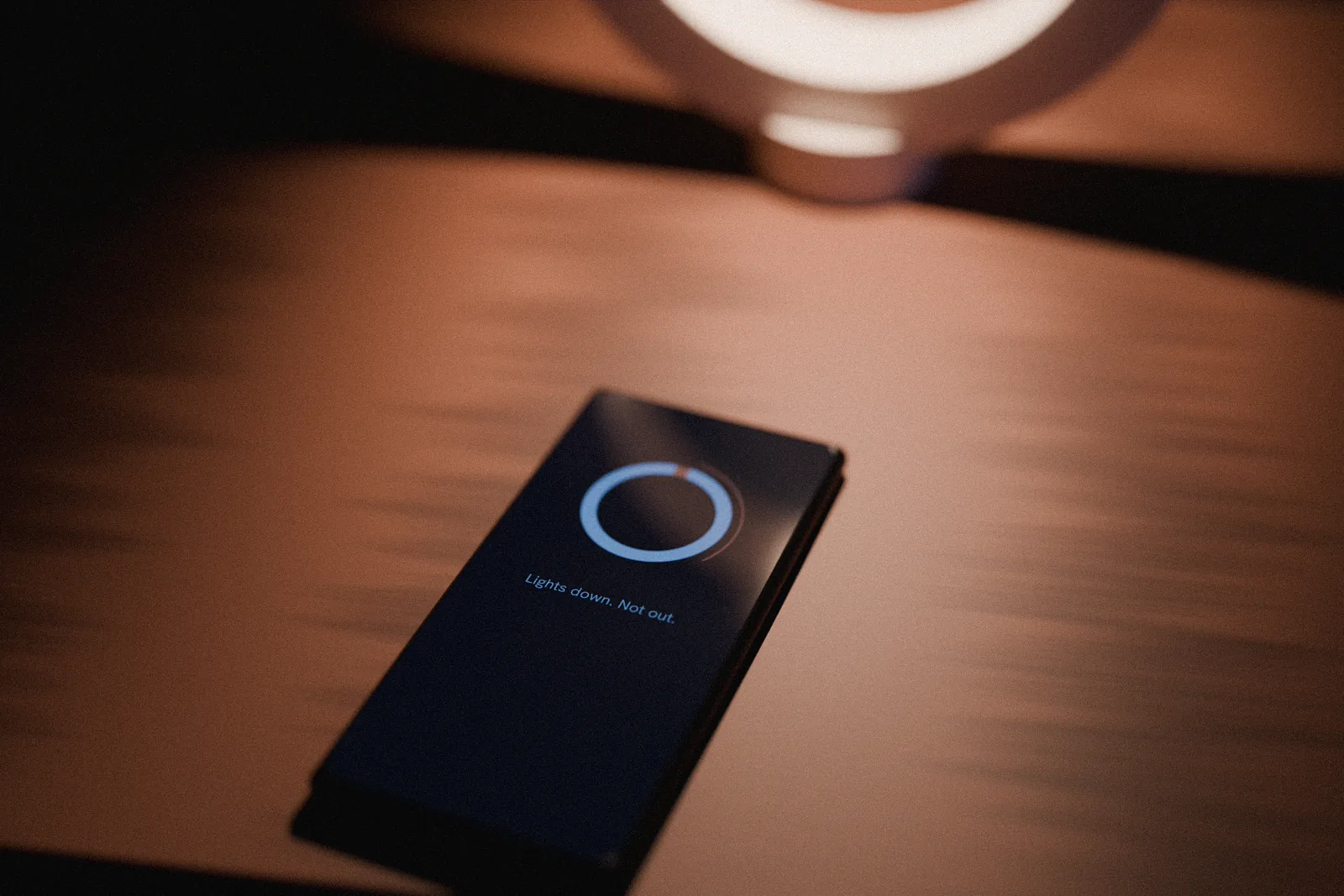 A phone lying on the bedside table showing the NOCT app: a dark screen, the ring as a dial and the line Lights down. Not out.