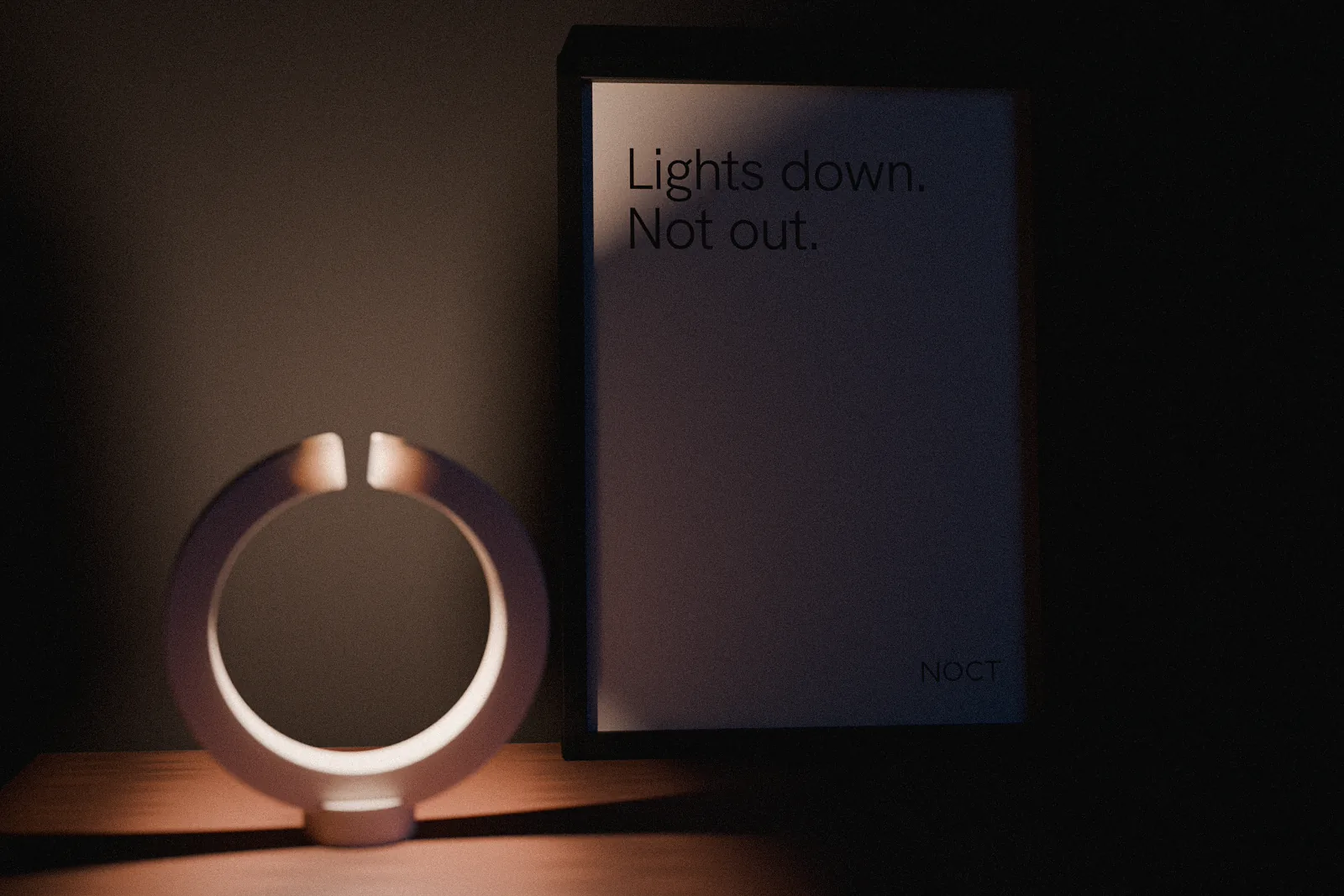 A framed NOCT print leaning on the wall beside the lamp: Lights down. Not out. set large, the wordmark small in the corner.