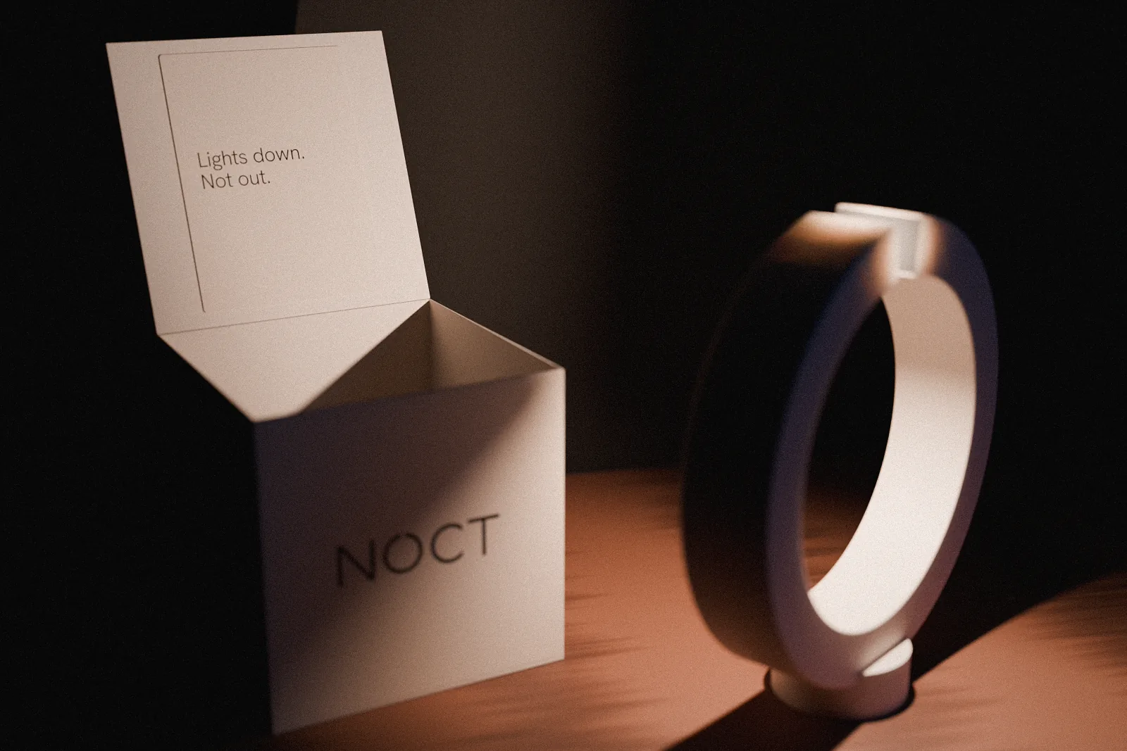 The NOCT box open on the table, its lid tipped back with a bone insert card reading Lights down. Not out., the lamp glowing beside it.