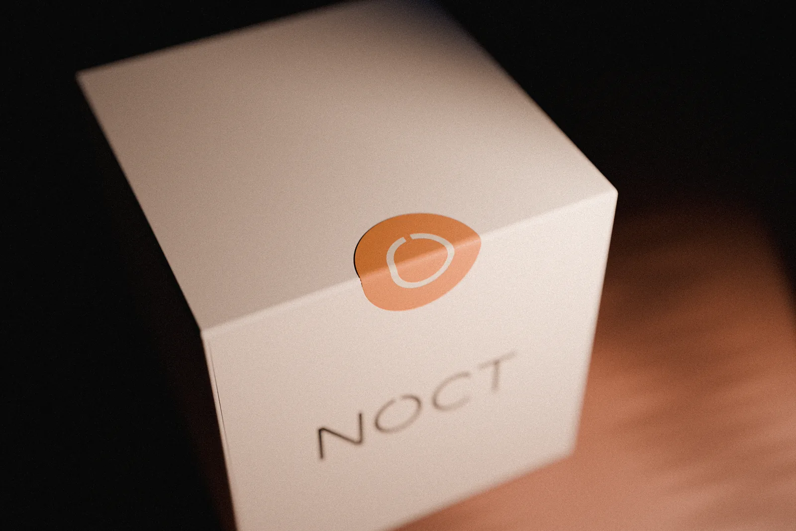 The closed NOCT box seen from above at an angle, a round ember sticker carrying the mark in bone folded over the lid’s front edge.