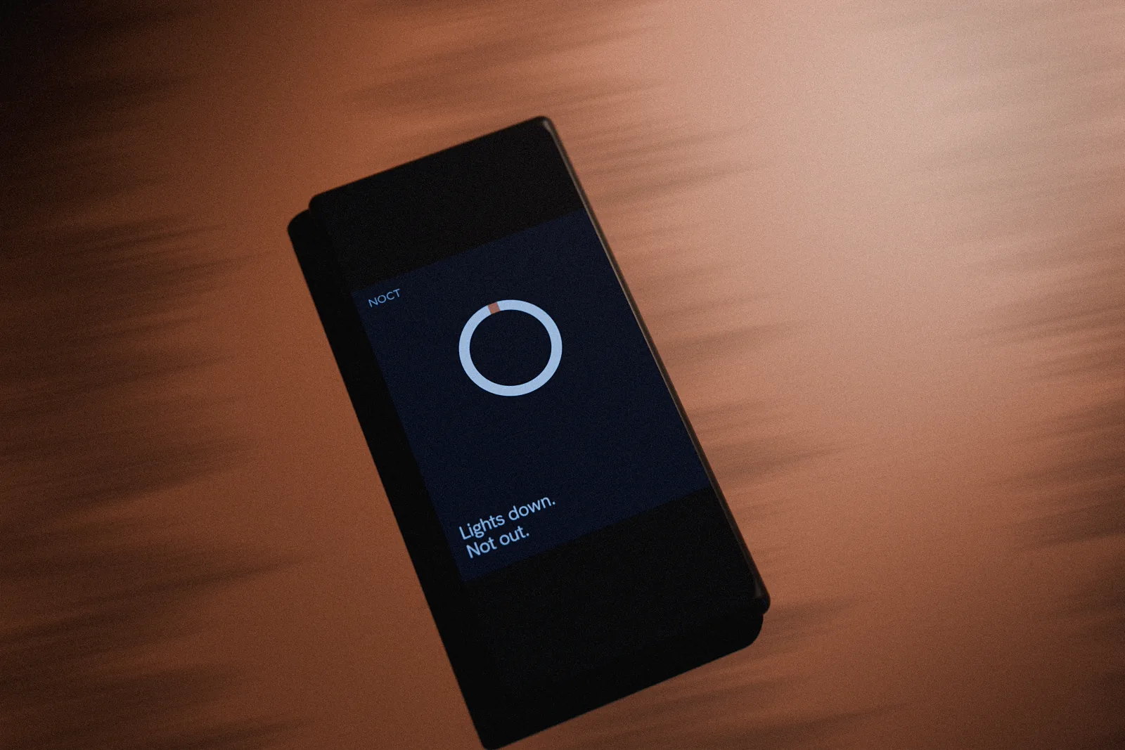 The phone on the table showing a NOCT post: the ring on a night background with the line Lights down. Not out. beneath it.