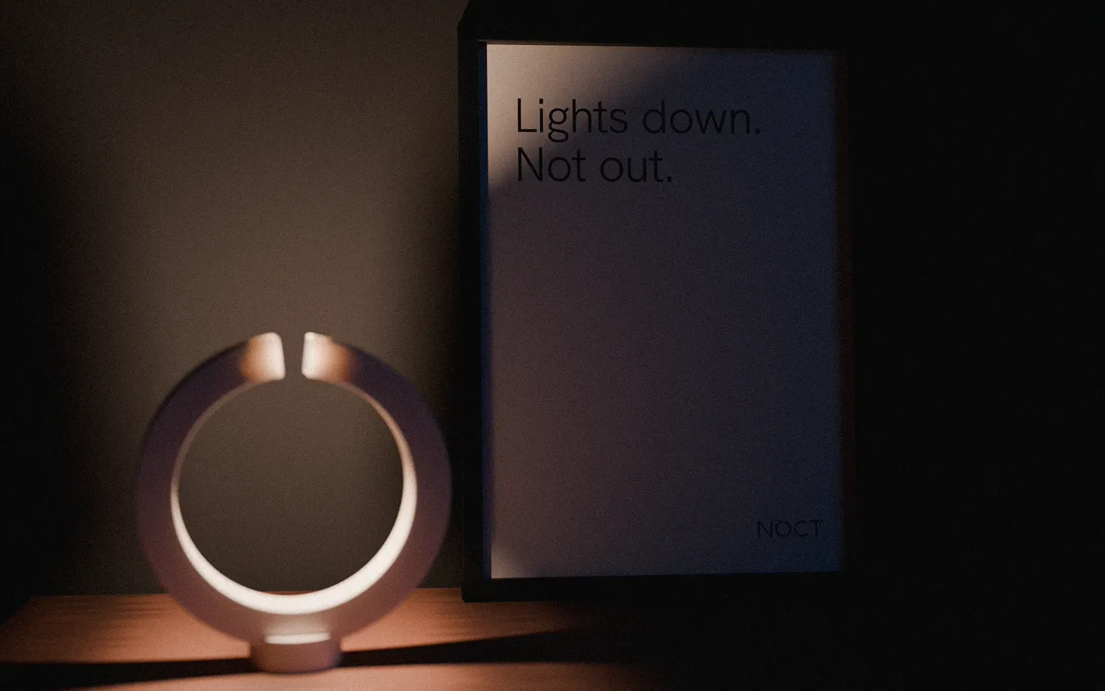 The NOCT lamp lit in the dark, a copper ring standing on its base beside the boxed product
